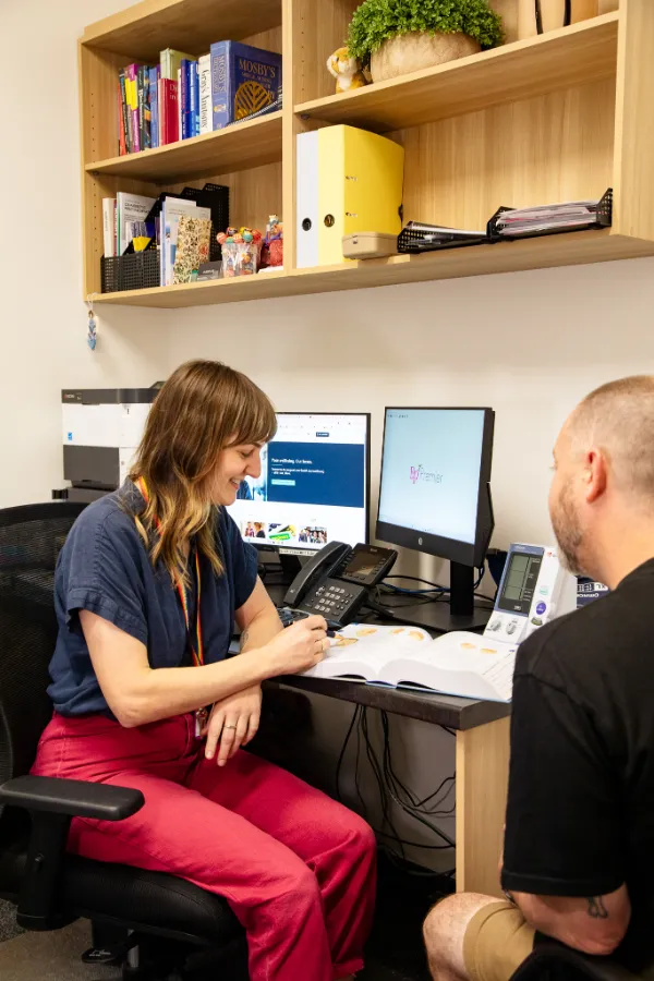 A woman sitting at a desk with a man, reviewing documents and using a computer in an office setting.