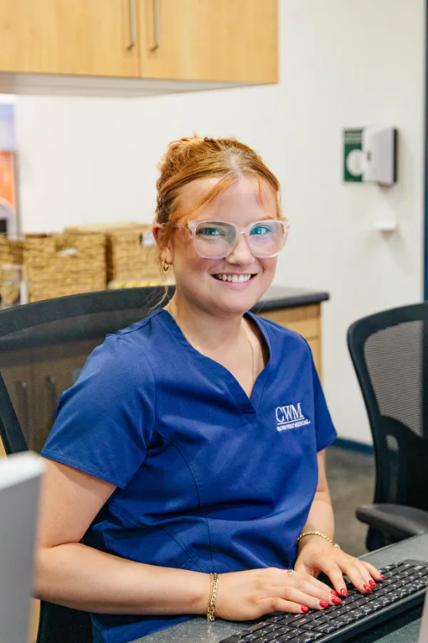 Smiling woman with red hair and glasses in blue medical scrubs typing on a keyboard at a desk in an office.