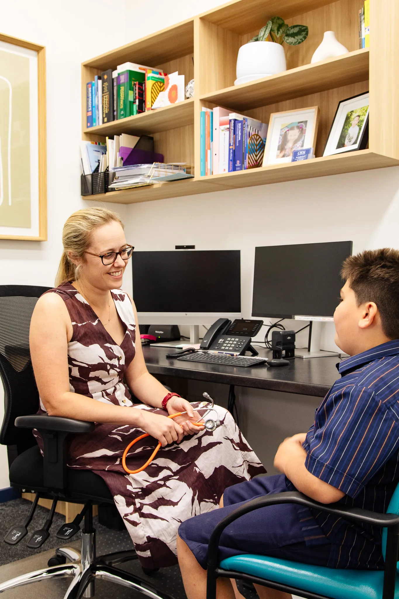 Female doctor with glasses and floral dress smiling and holding a stethoscope while talking to a boy in a blue striped shirt in a medical office.