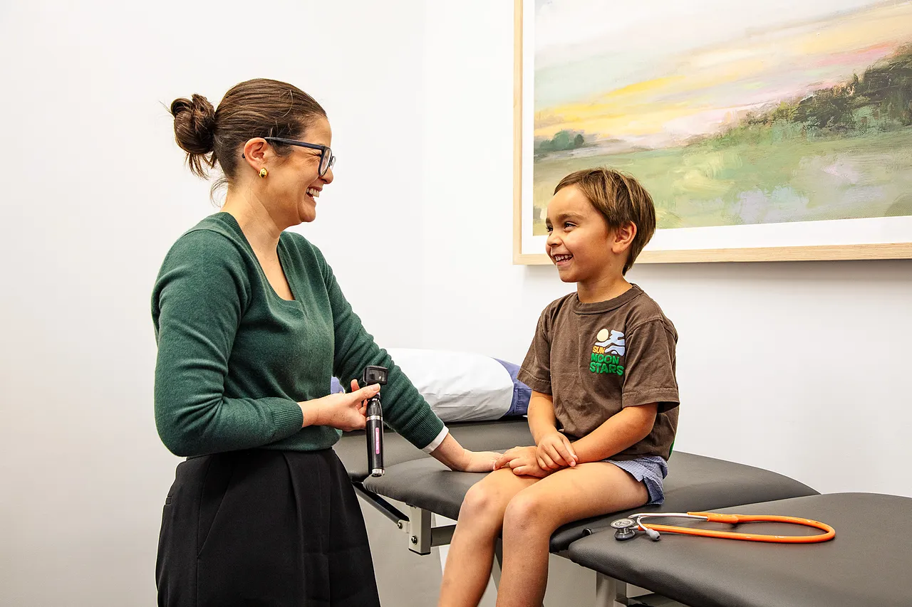 Smiling female doctor holding an otoscope while interacting with a happy young boy sitting on an examination table.