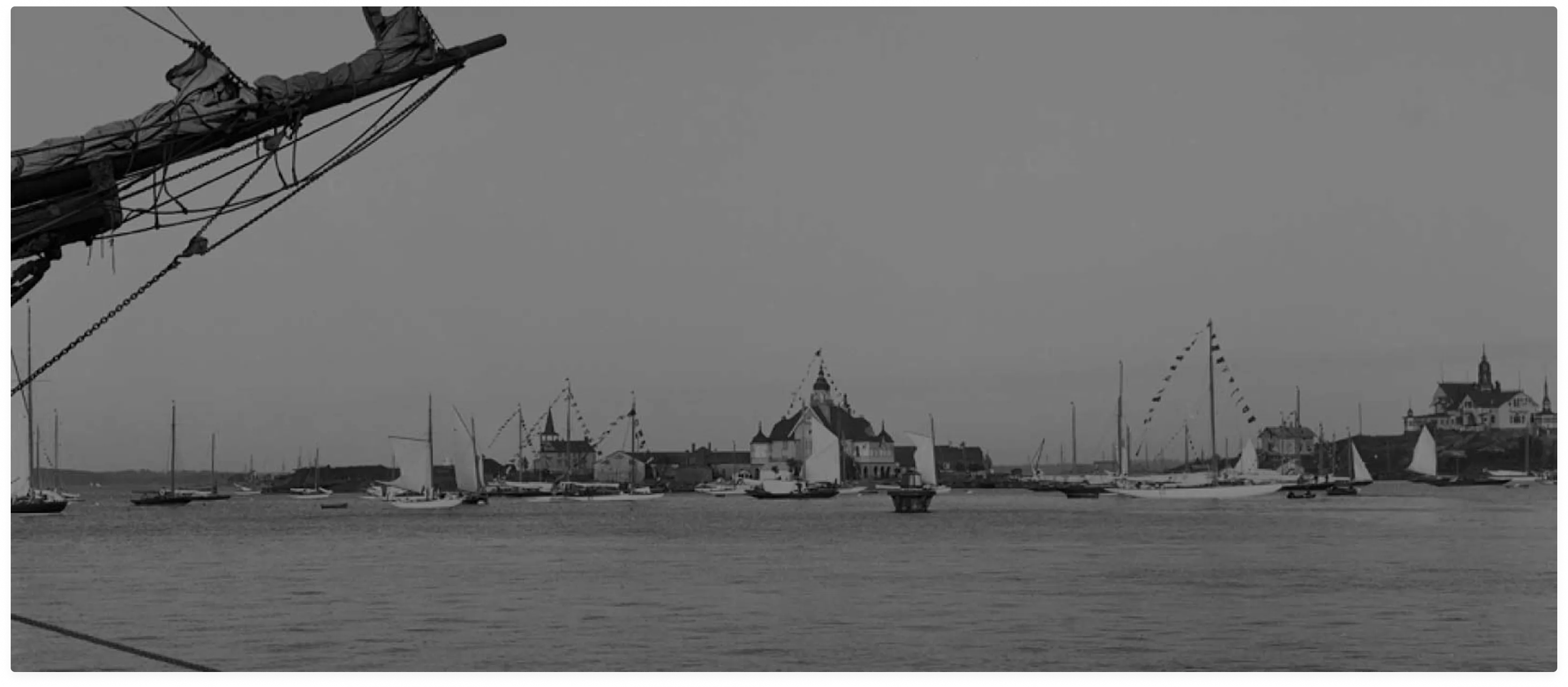 Svartvitt foto av en hamn med segelbåtar och byggnader med flaggor i bakgrunden.