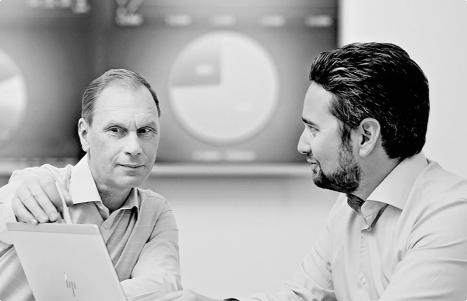 Två affärsmän i vit skjorta diskuterar framför en skärm med cirkeldiagram.