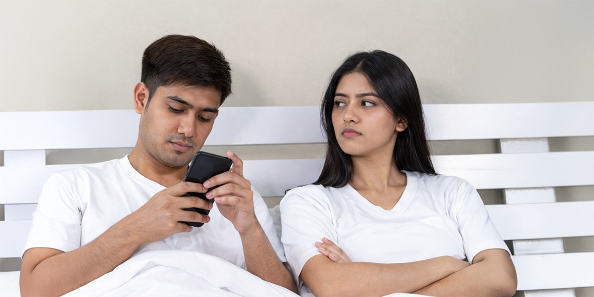 Couple sitting in bed with man using phone and woman looking concerned, representing relationship stress linked to erectile dysfunction and diabetes