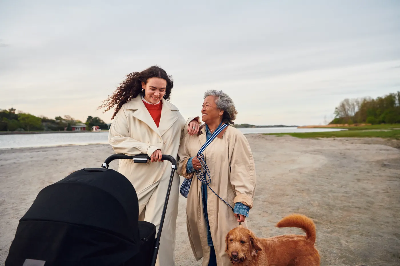 Två kvinnor i beiga kappor promenerar vid en strand, den ena skjuter en barnvagn och den andra håller i en brun hund i koppel.