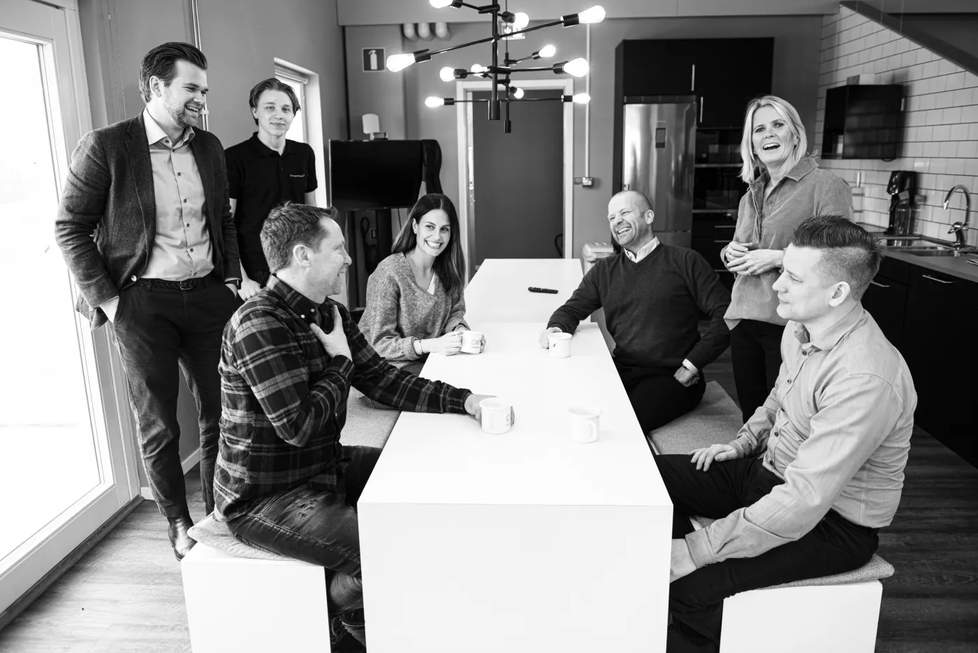 Sju personer sitter och står runt ett bord i ett modernt kontorsköksrum och pratar och skrattar under en fikapaus.