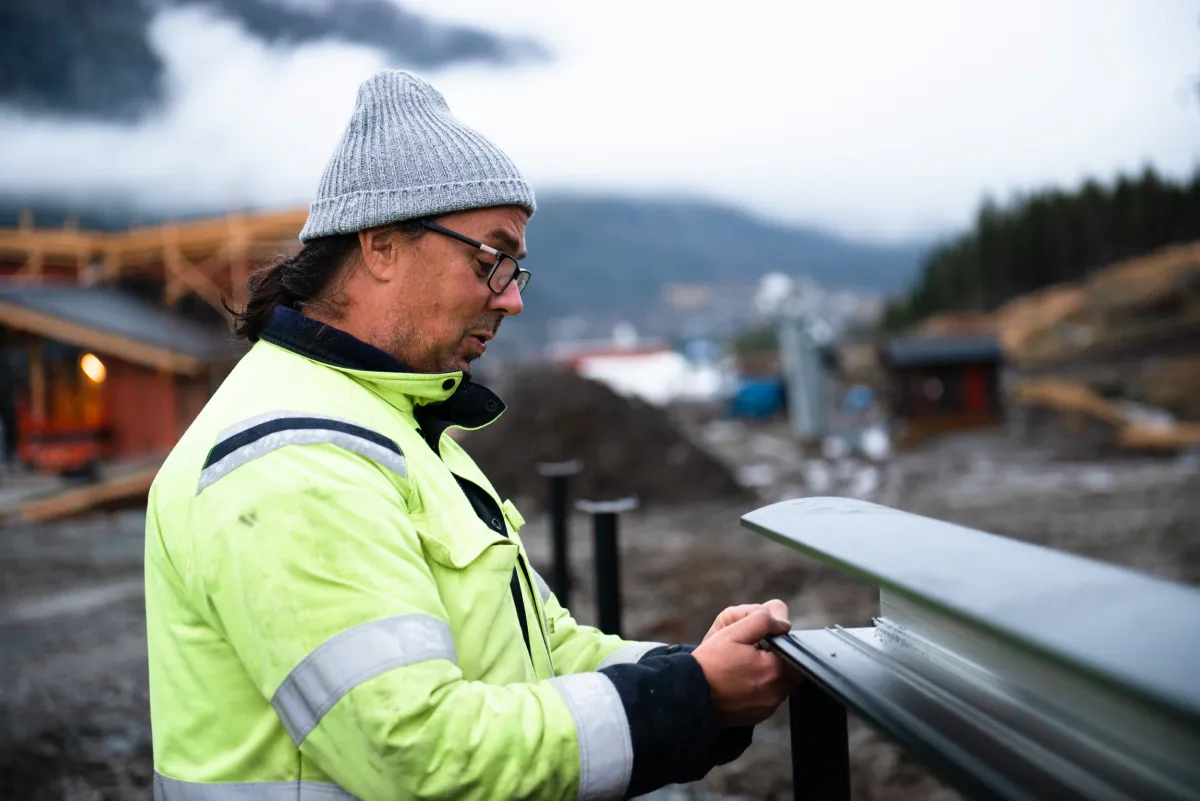 En arbetare med grå mössa och reflexjacka installerar en laddstation utomhus i ett bergigt område.