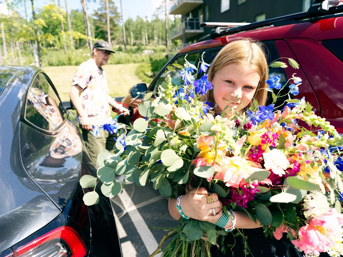 Flicka håller en stor bukett färgglada blommor nära en röd bil medan en man med keps står vid en svart bil i bakgrunden.