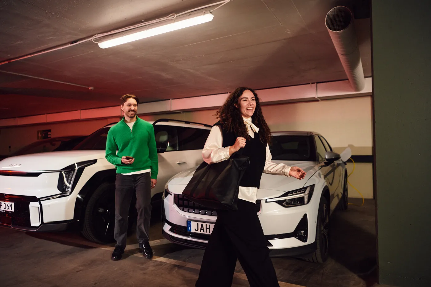 Kvinna går glad i ett garage framför två vita elbilar, en man i grön tröja står bredvid en av bilarna.