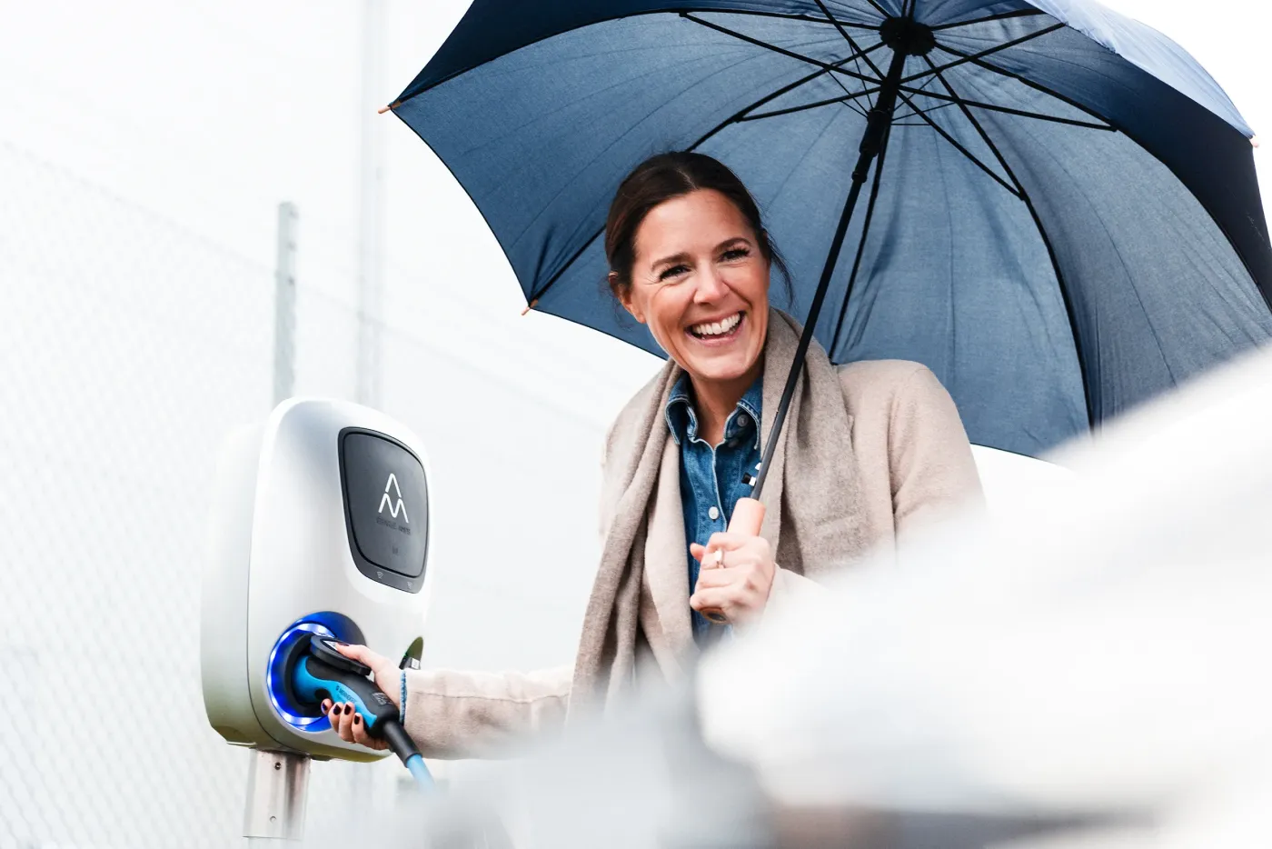 Leende kvinna laddar elbil under ett paraply på en regnig dag.