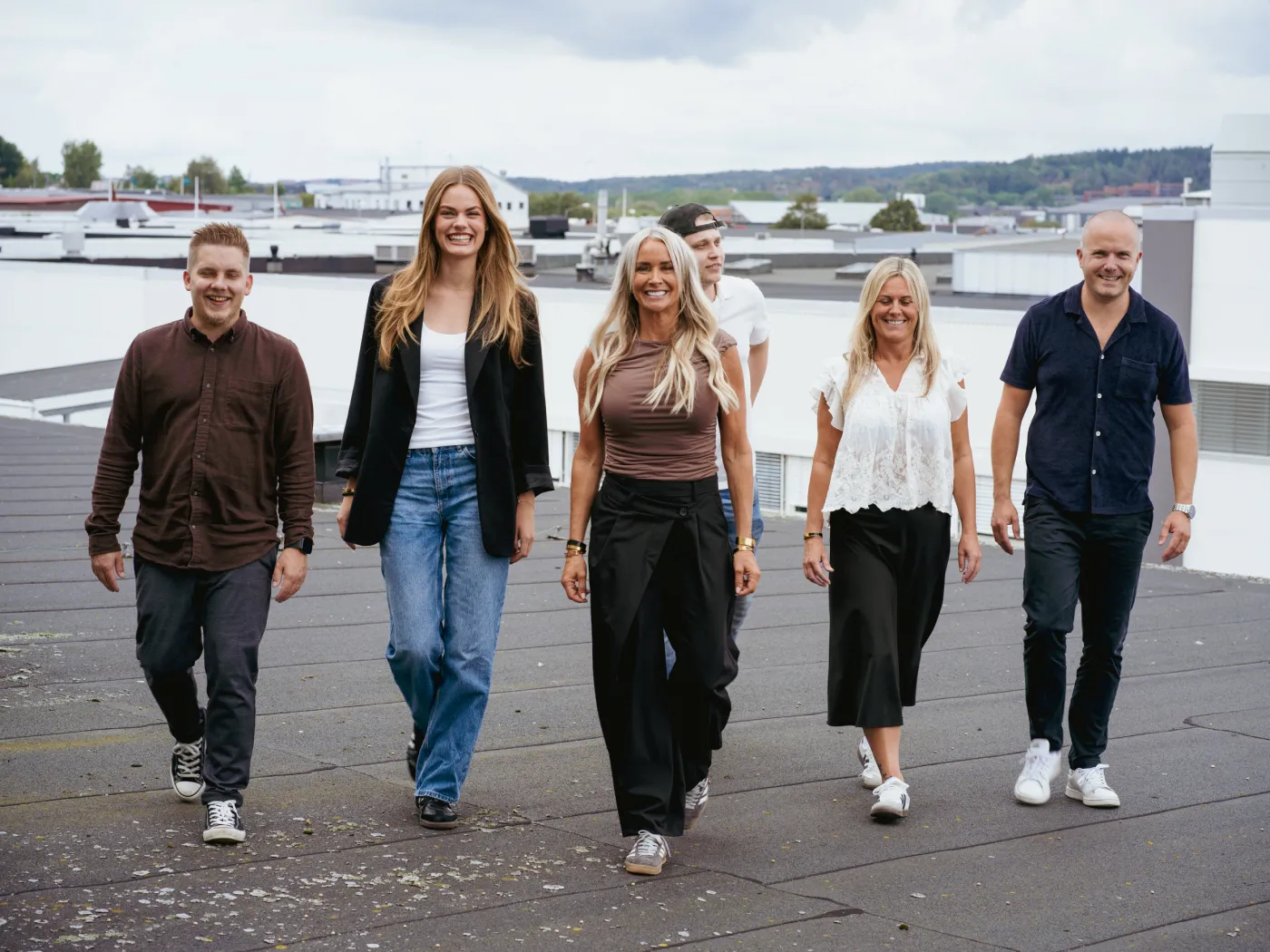 Sex leende personer som går mot kameran på ett tak med stadssilhuett i bakgrunden.