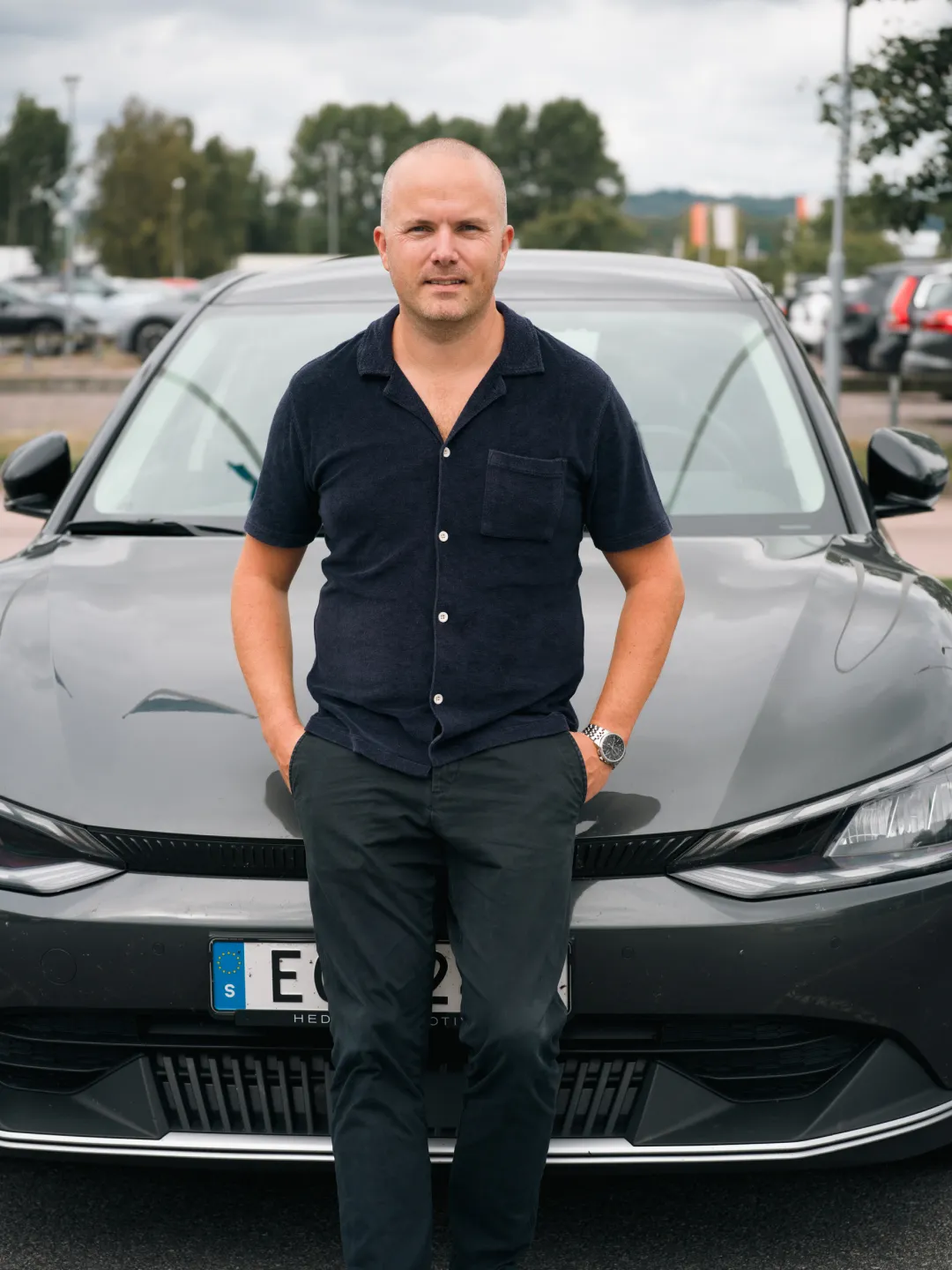 Man står framför en mörkgrå bil i en parkeringsplats med händerna i byxfickorna.