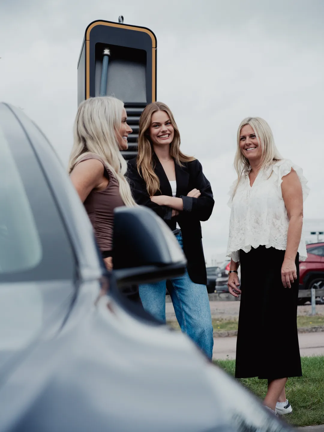 Tre leende kvinnor står vid en elbilsladdningsstation bredvid en parkerad bil.