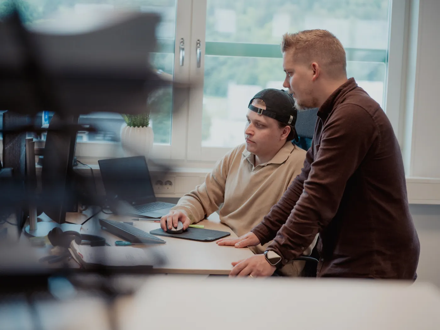 Två män samarbetar framför en dator i ett ljust kontorsrum med stort fönster.