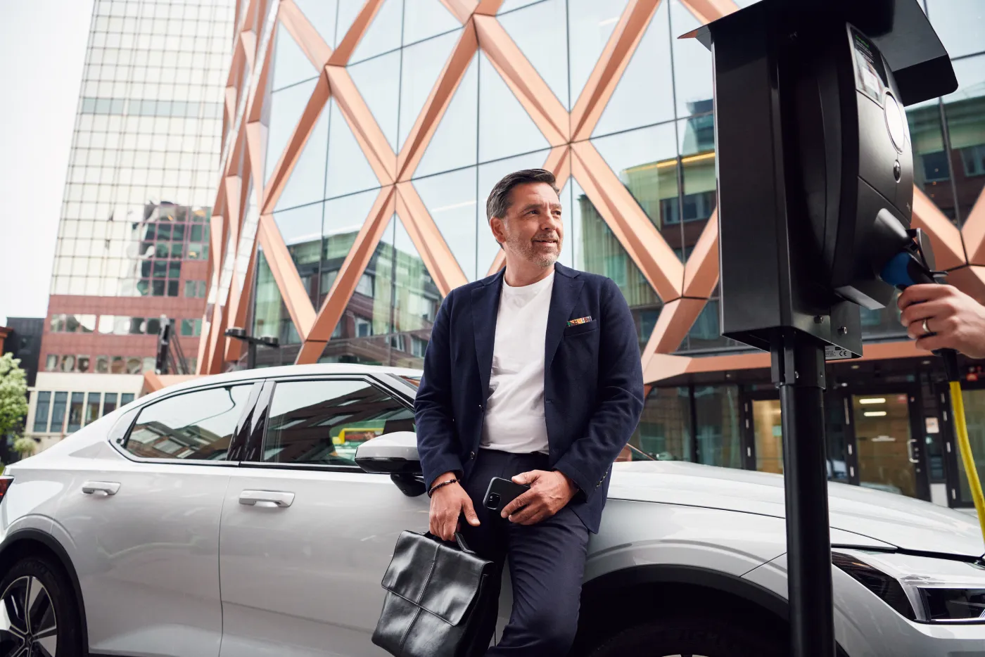 Man in business attire lutar sig mot en vit elbil som laddas vid en laddstation framför en modern glasbyggnad med geometriska mönster.