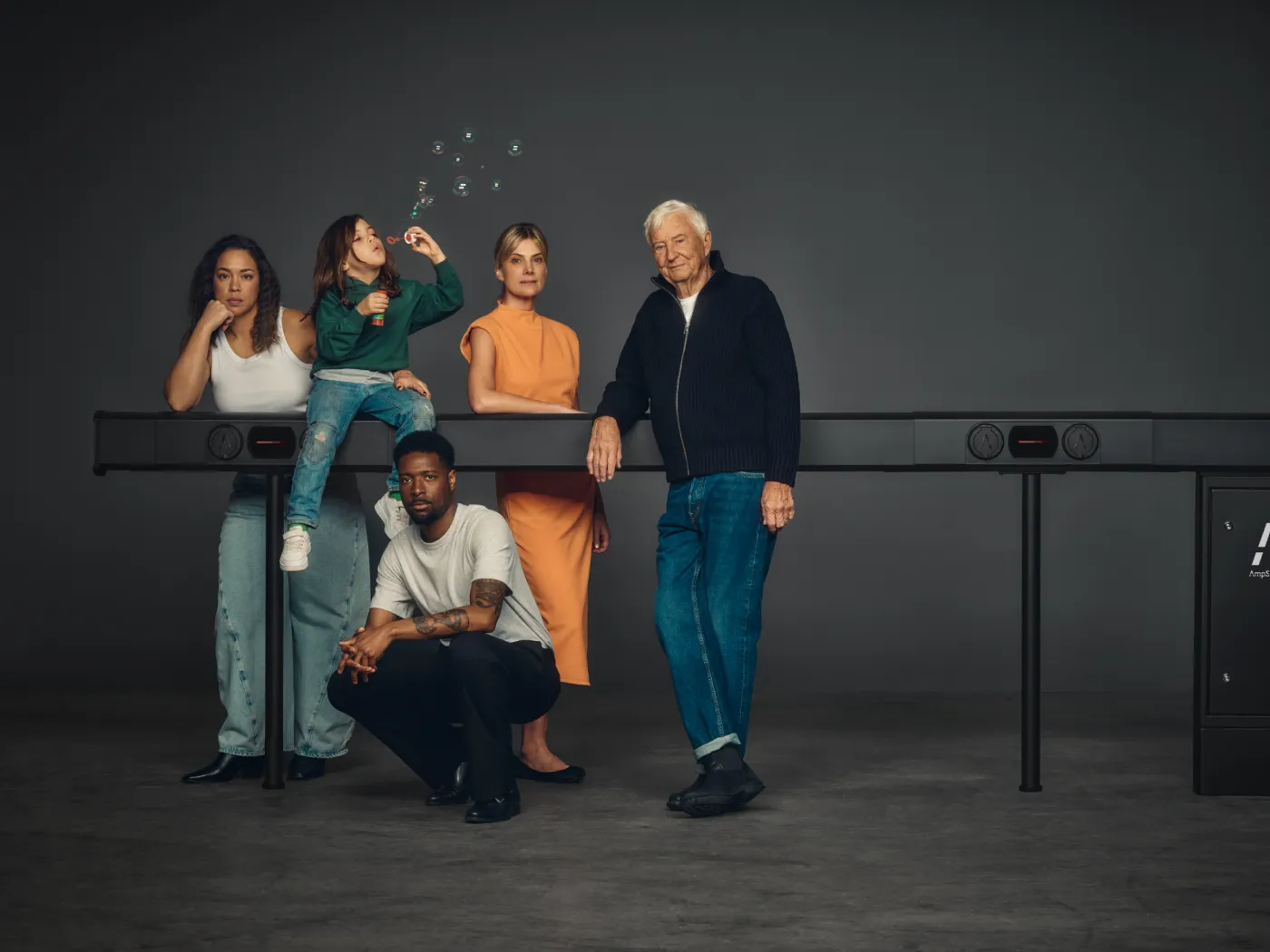 En grupp på fem personer i olika åldrar poserar tillsammans vid en lång svart enhet mot en mörk bakgrund.