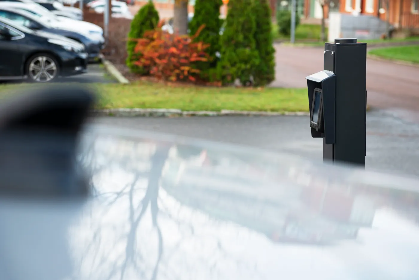 Utomhus betalterminal för elbilsladdning i parkeringsområde med bilar i bakgrunden.