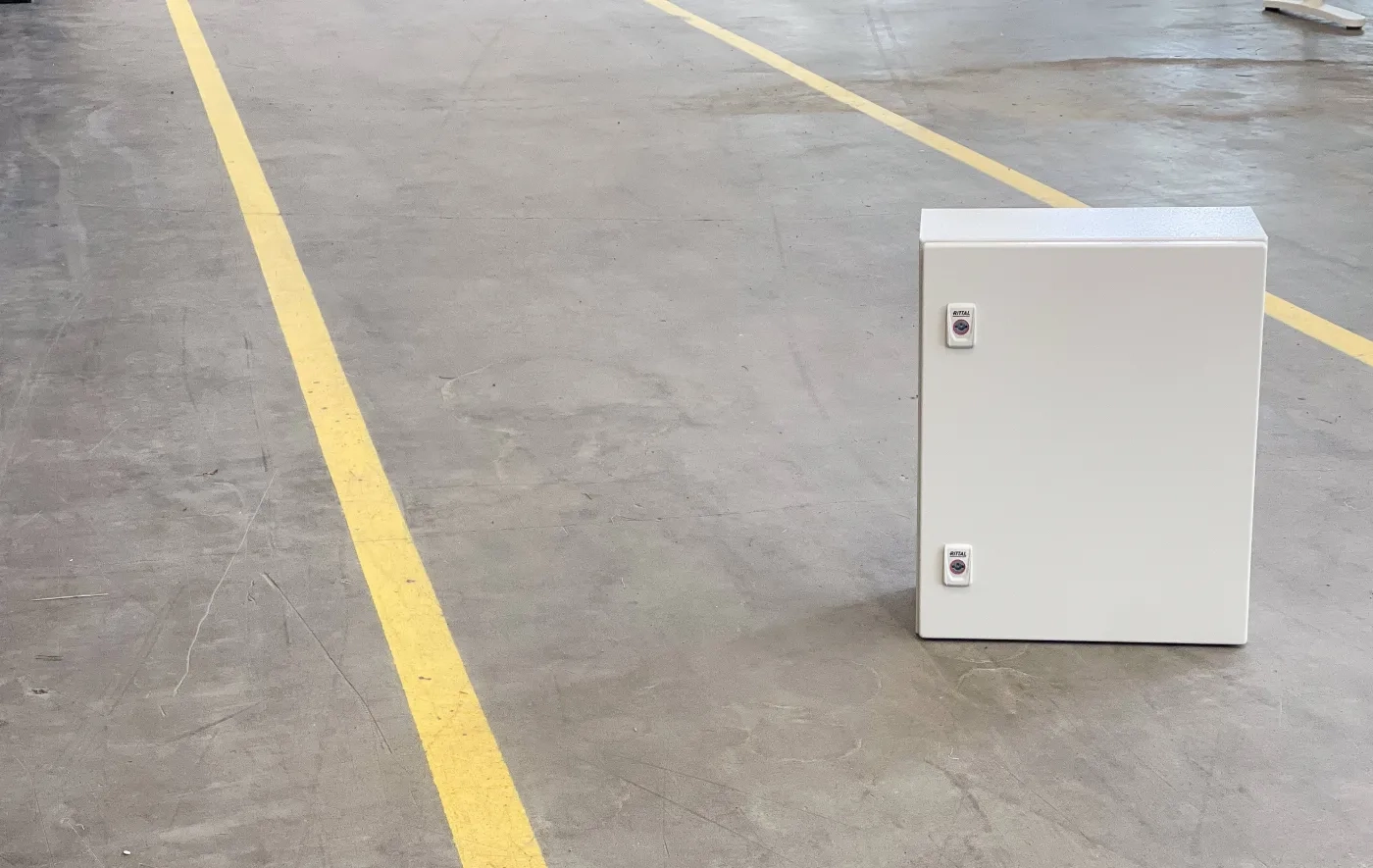 Vit elektrisk kopplingsbox placerad på betonggolv med gula linjer i ett industrilager.