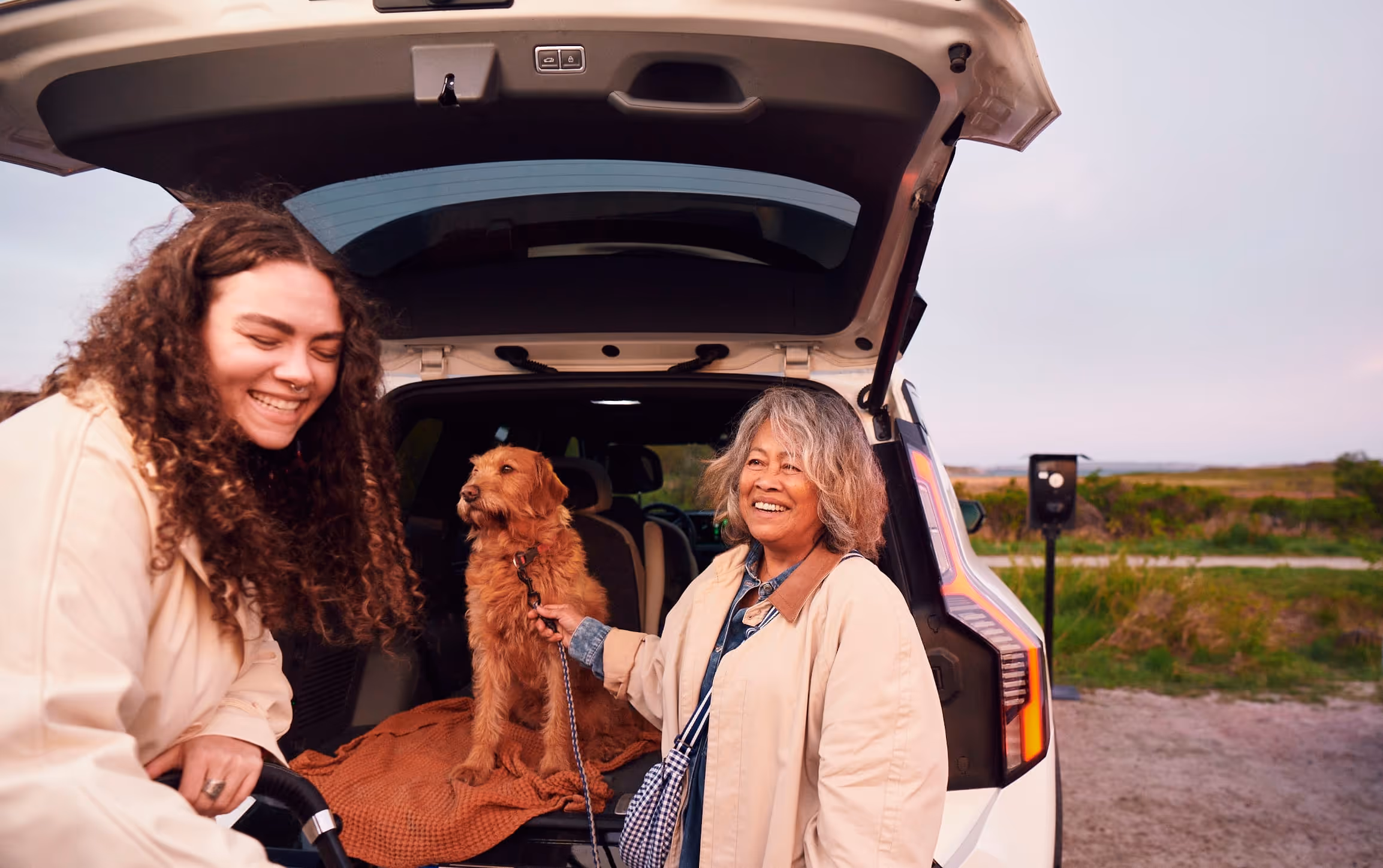 Två kvinnor vid en öppen bilbaklucka med en brun hund som sitter på en filt inuti bilen.