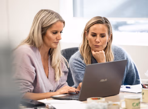 Två kvinnor sitter vid ett bord och samarbetar medan de tittar på skärmen på en bärbar dator.