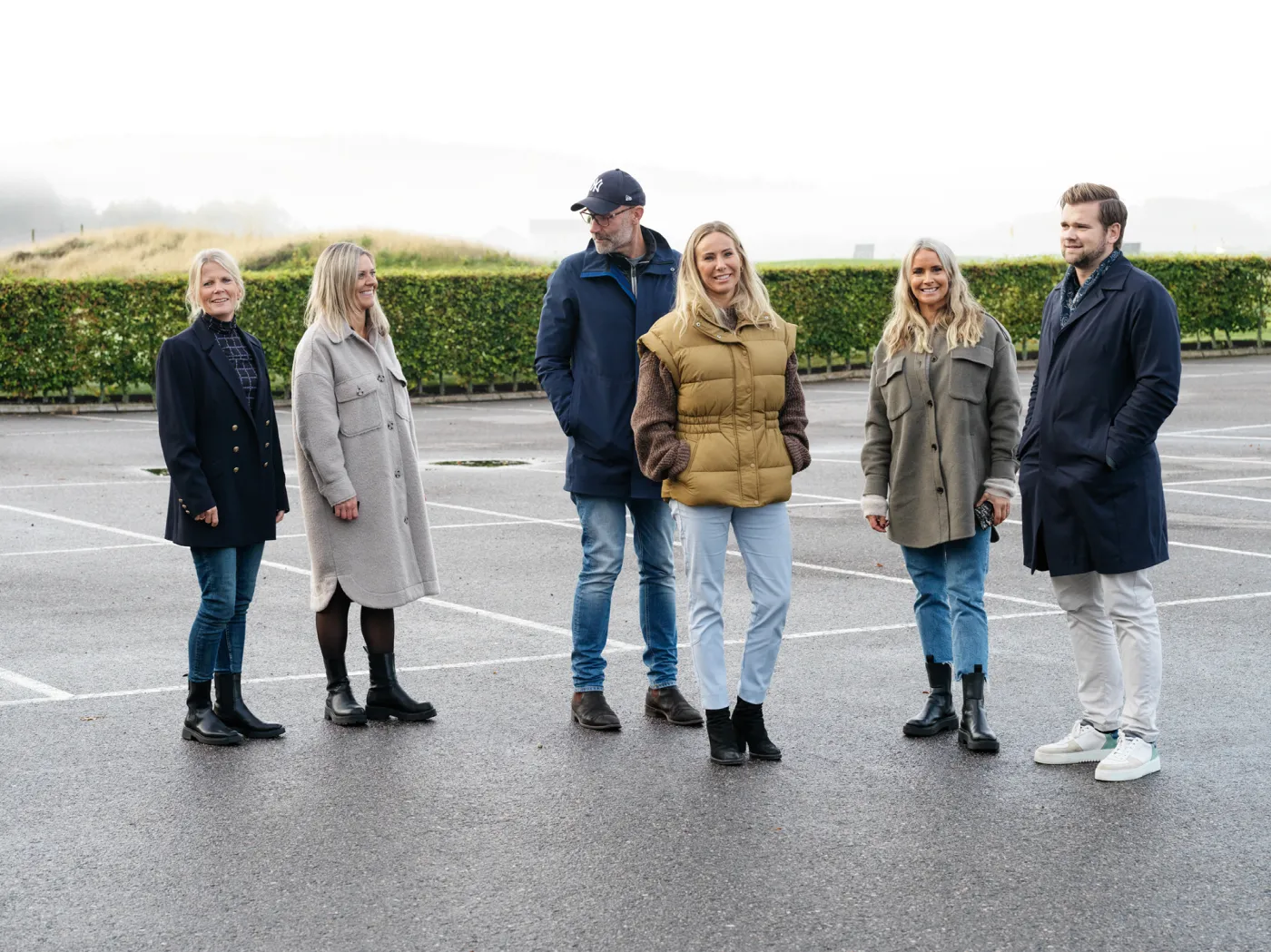 En grupp på sex personer står utomhus på en parkeringsplats med grön häck i bakgrunden, klädda i höstkläder och ler mot kameran.