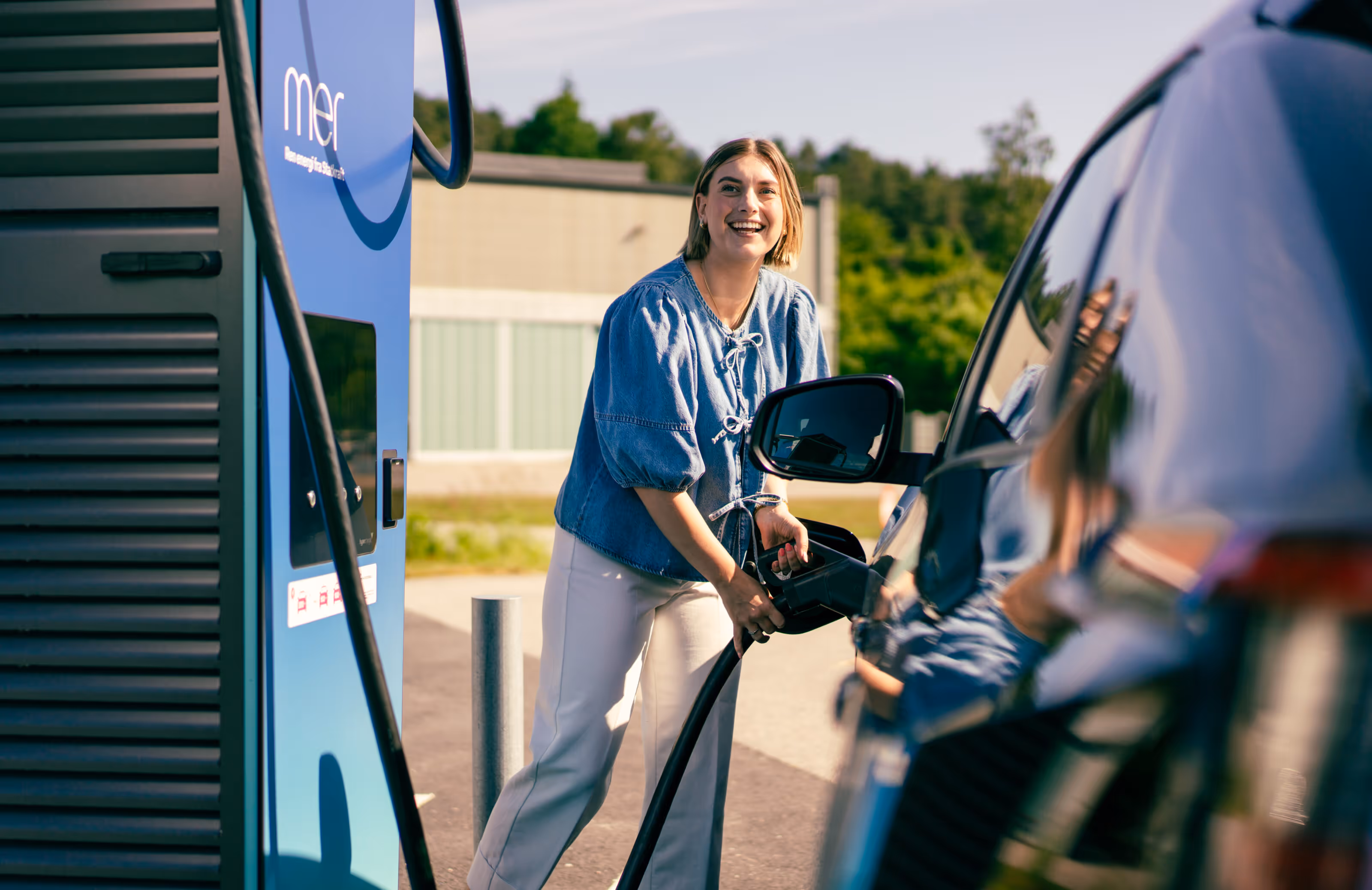 Leende kvinna som tankar sin bil vid en elbilsladdningsstation på en solig dag.