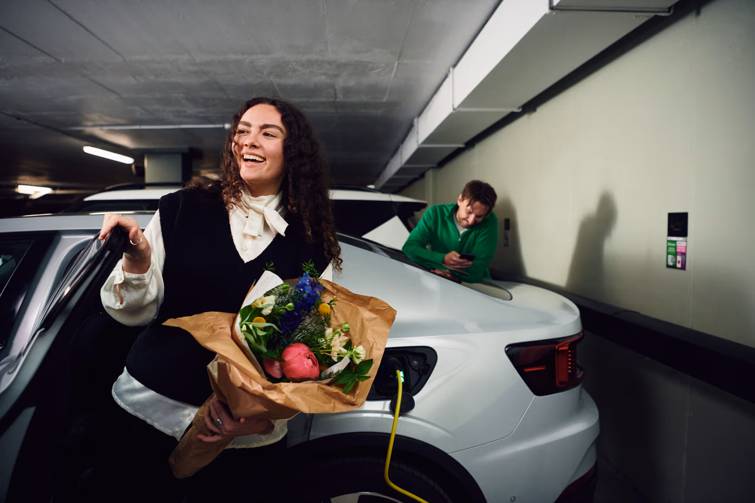 Leende kvinna håller en bukett blommor medan en man laddar en vit elbil i ett parkeringsgarage.