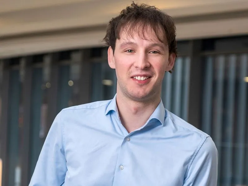 Smiling young man with tousled brown hair wearing a light blue button-up shirt in a modern office setting.