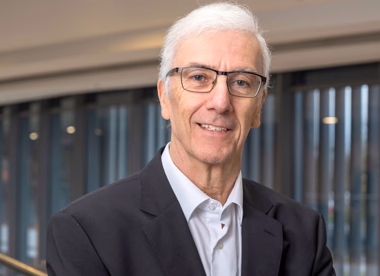 Smiling elderly man with white hair and glasses wearing a black suit jacket and white shirt indoors.