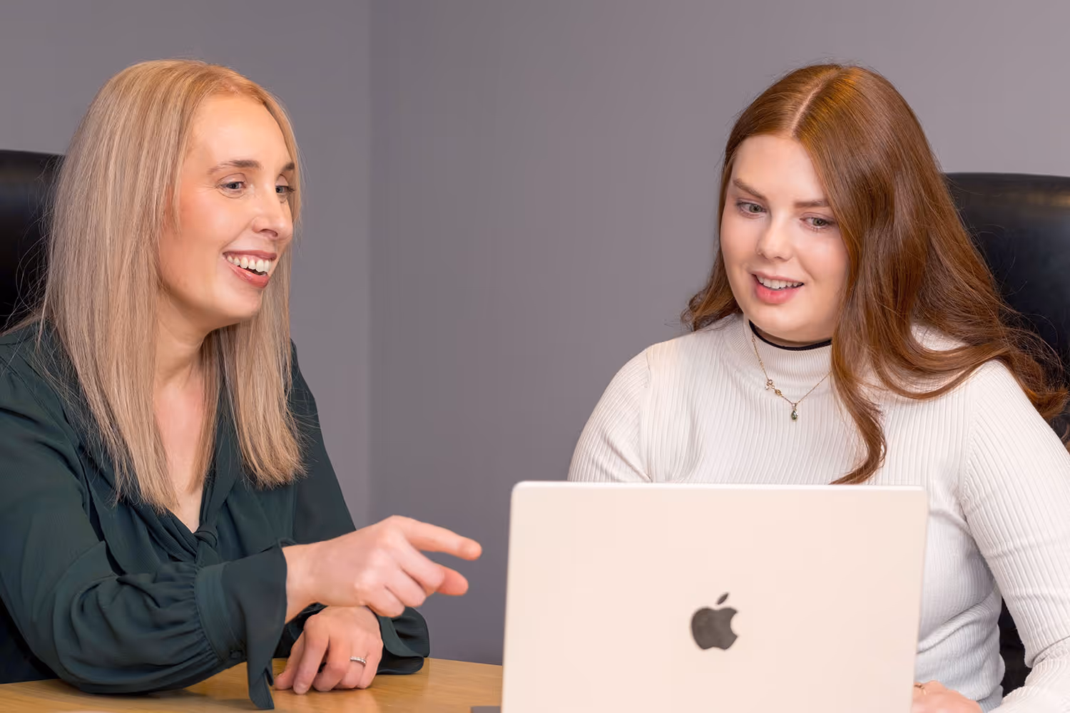 Two women sitting at a desk sharing a laptop, one pointing at the screen and both smiling.