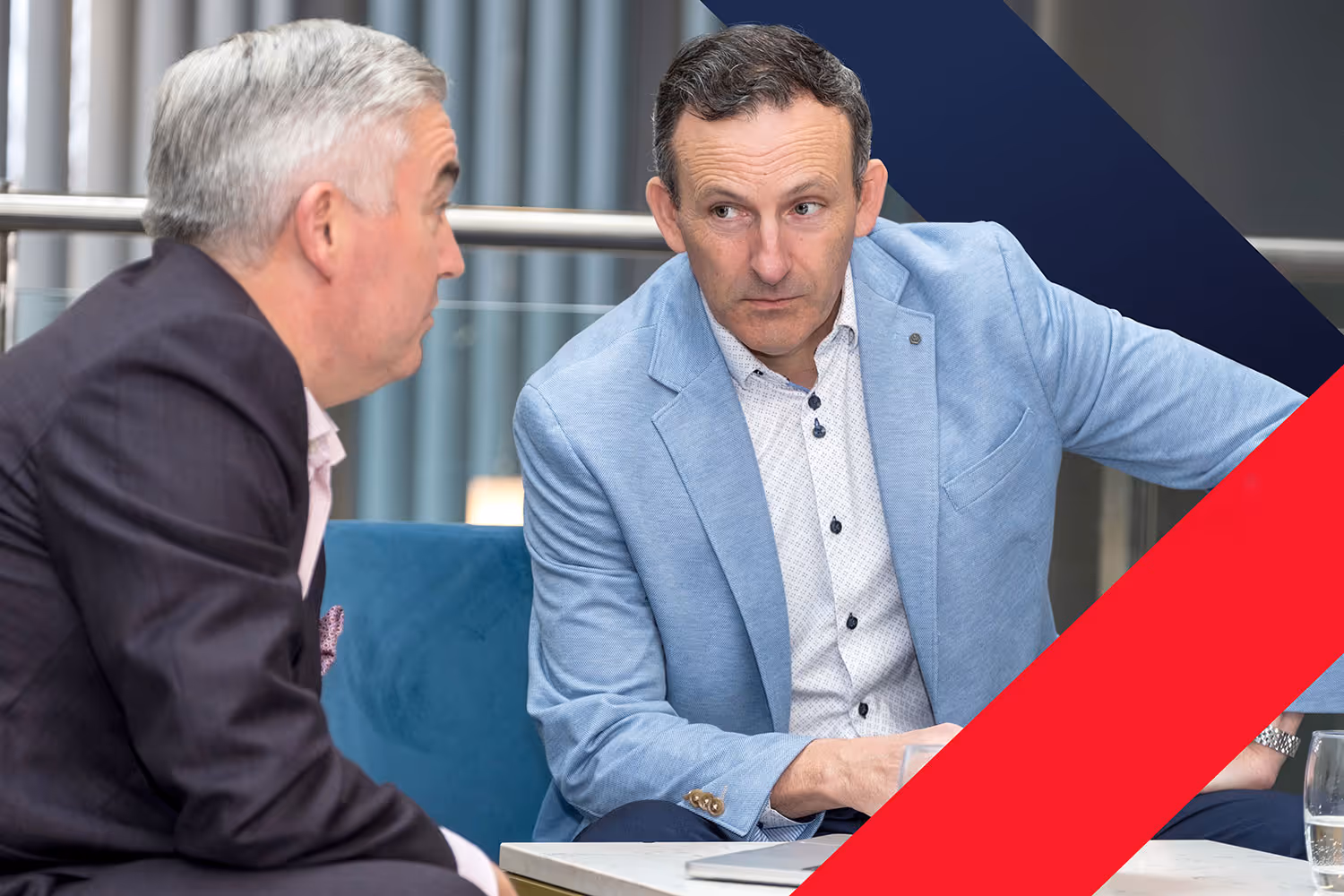 Two men in suits engaged in a serious conversation while sitting in a modern office lounge area.