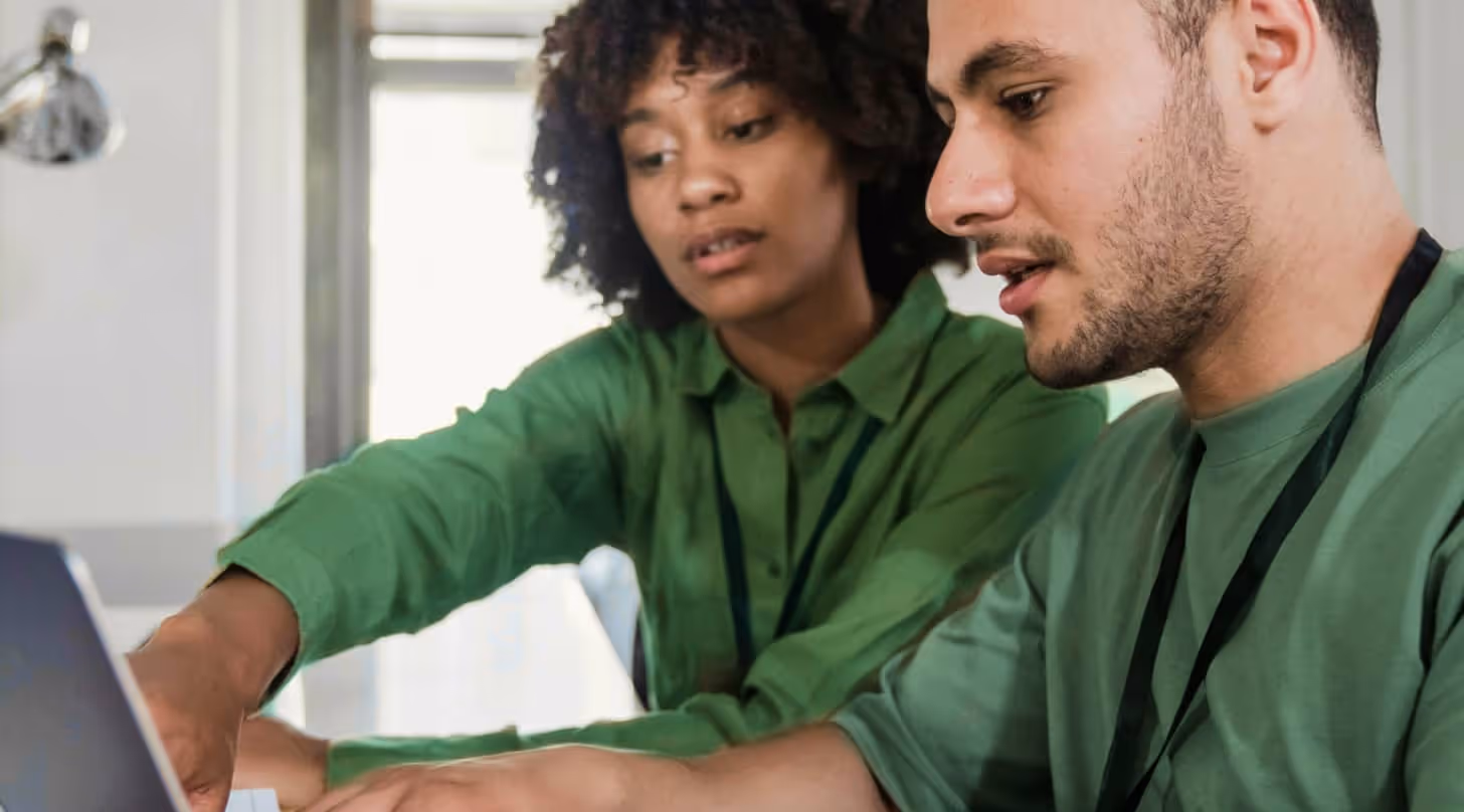 Two colleagues in green shirts collaborating and looking at a laptop screen.