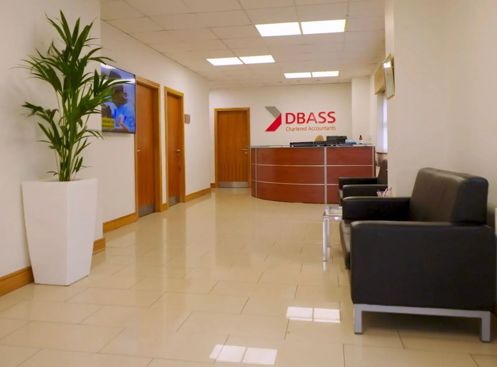 Modern reception area with wooden doors, brown curved reception desk with DBASS Chartered Accountants logo, black sofas, and a large potted plant.