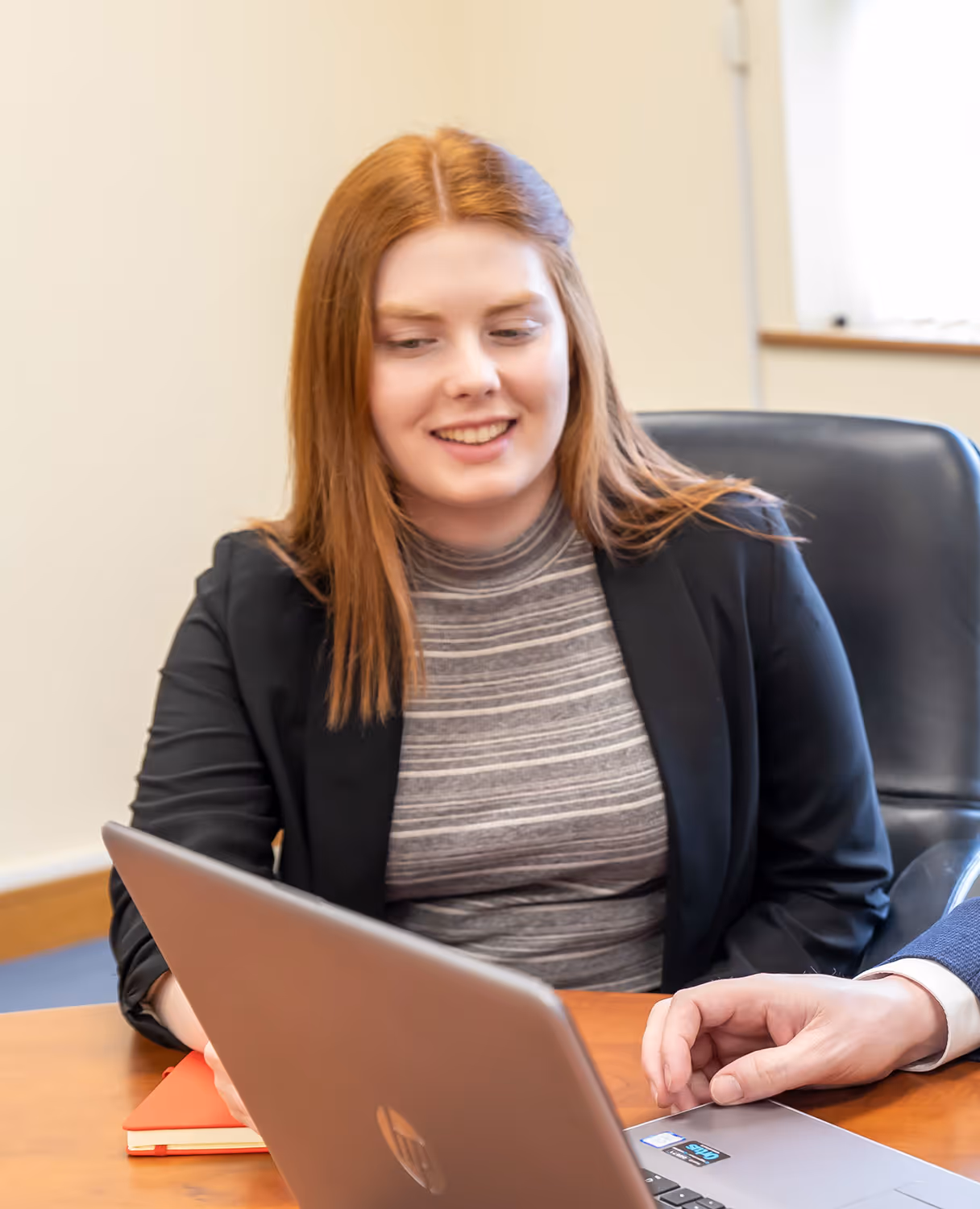 Smiling woman with red hair wearing a black blazer looking at a laptop screen while sitting at a table.