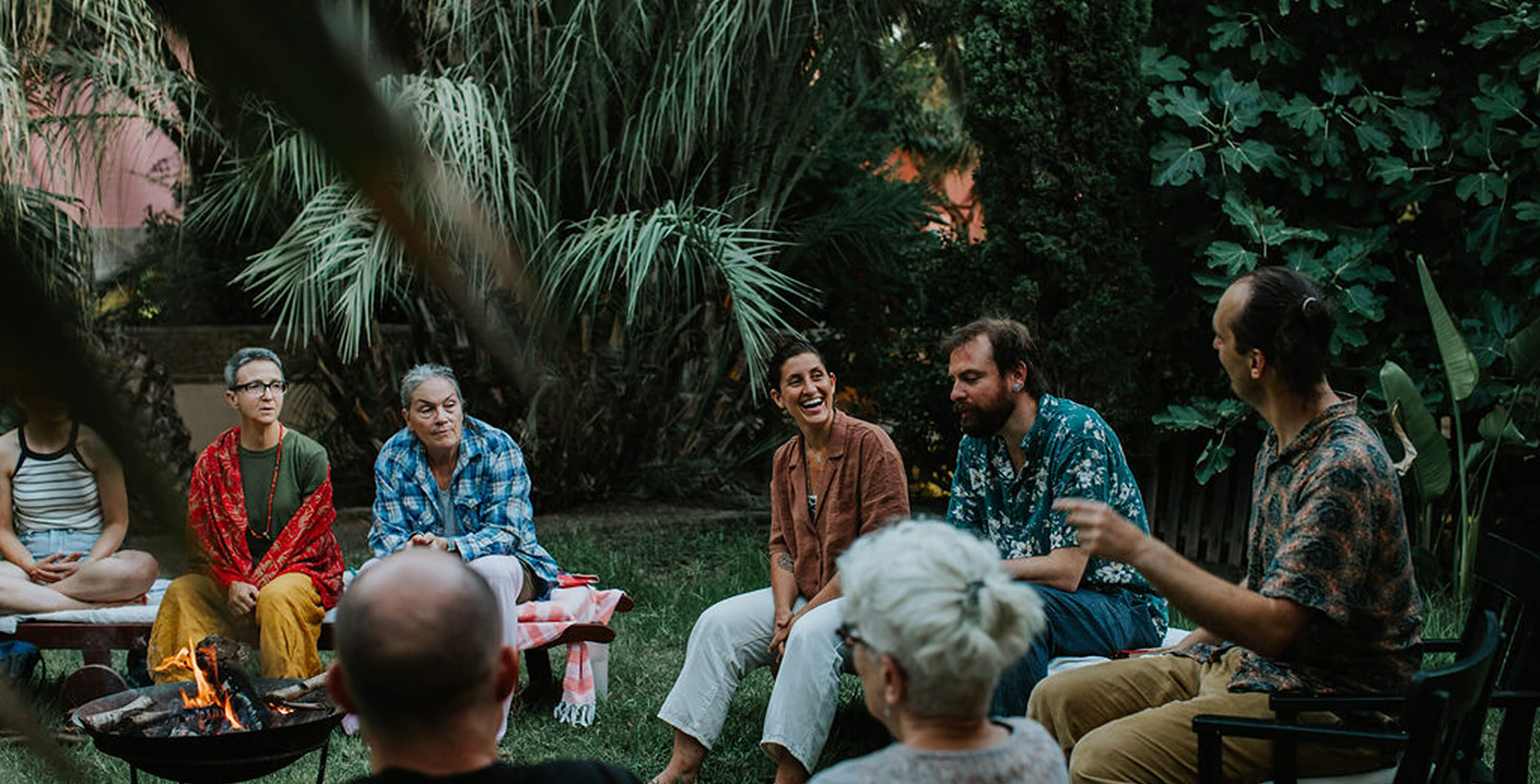 Group of adults sitting in a garden around a small fire pit, engaged in lively conversation.