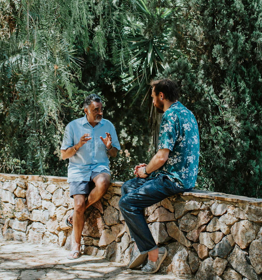 Two men having a conversation while sitting on a stone wall surrounded by lush greenery.