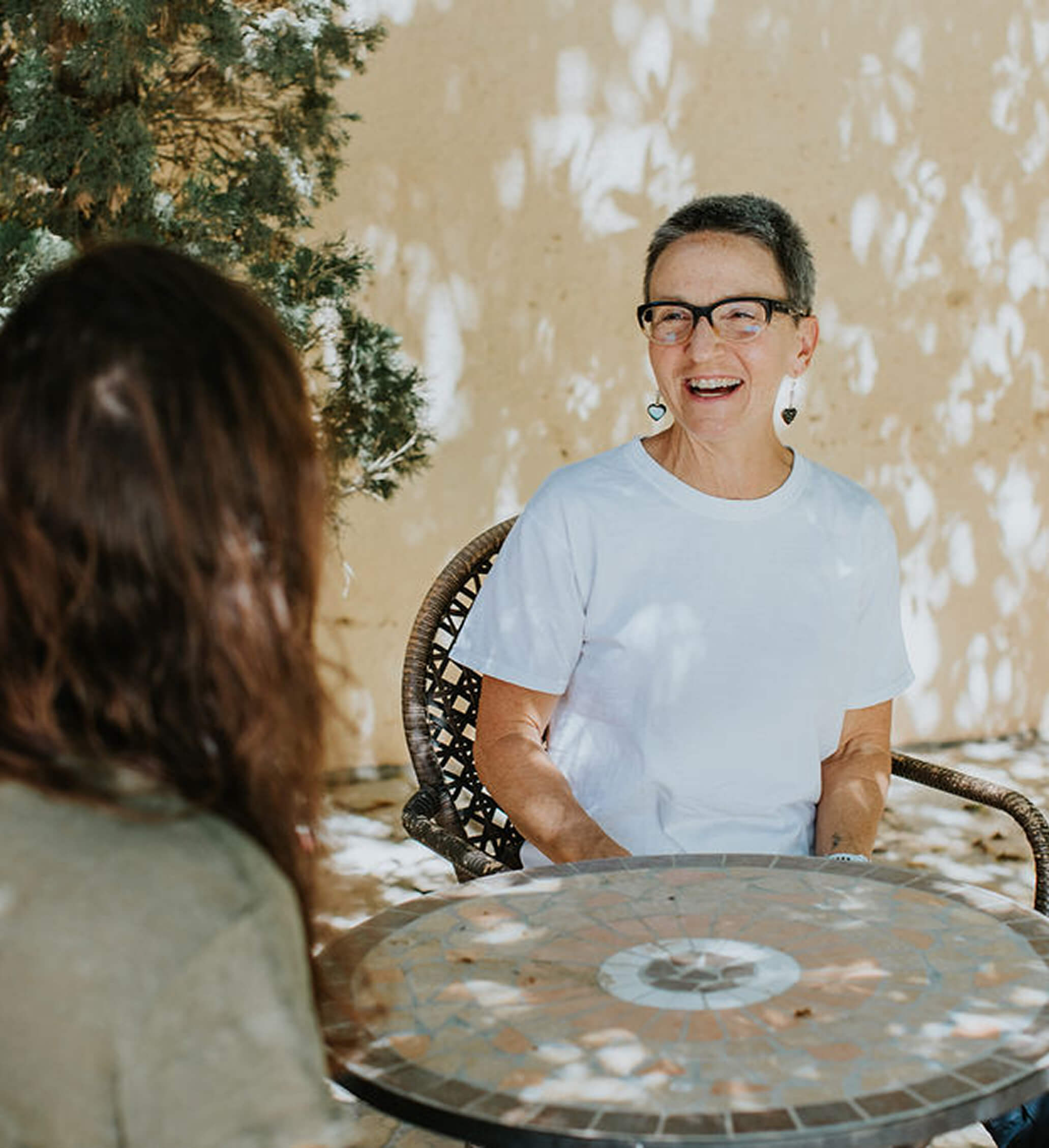 Two women sitting at a mosaic table outdoors, one woman with short gray hair and glasses smiling.
