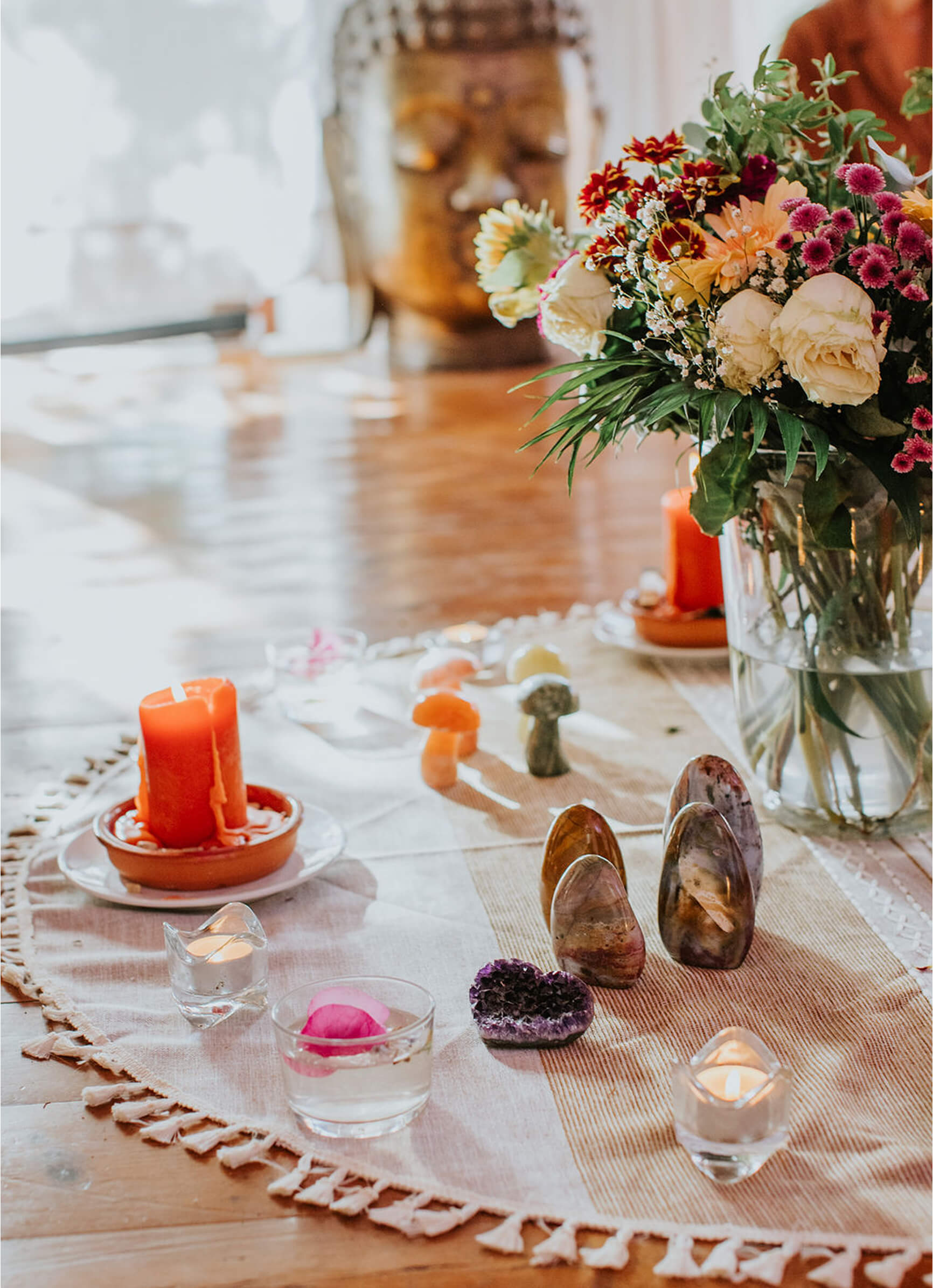 Table with lit candles, crystals, a glass of water with a rose petal, and a bouquet of colorful flowers with a blurred Buddha statue in the background.