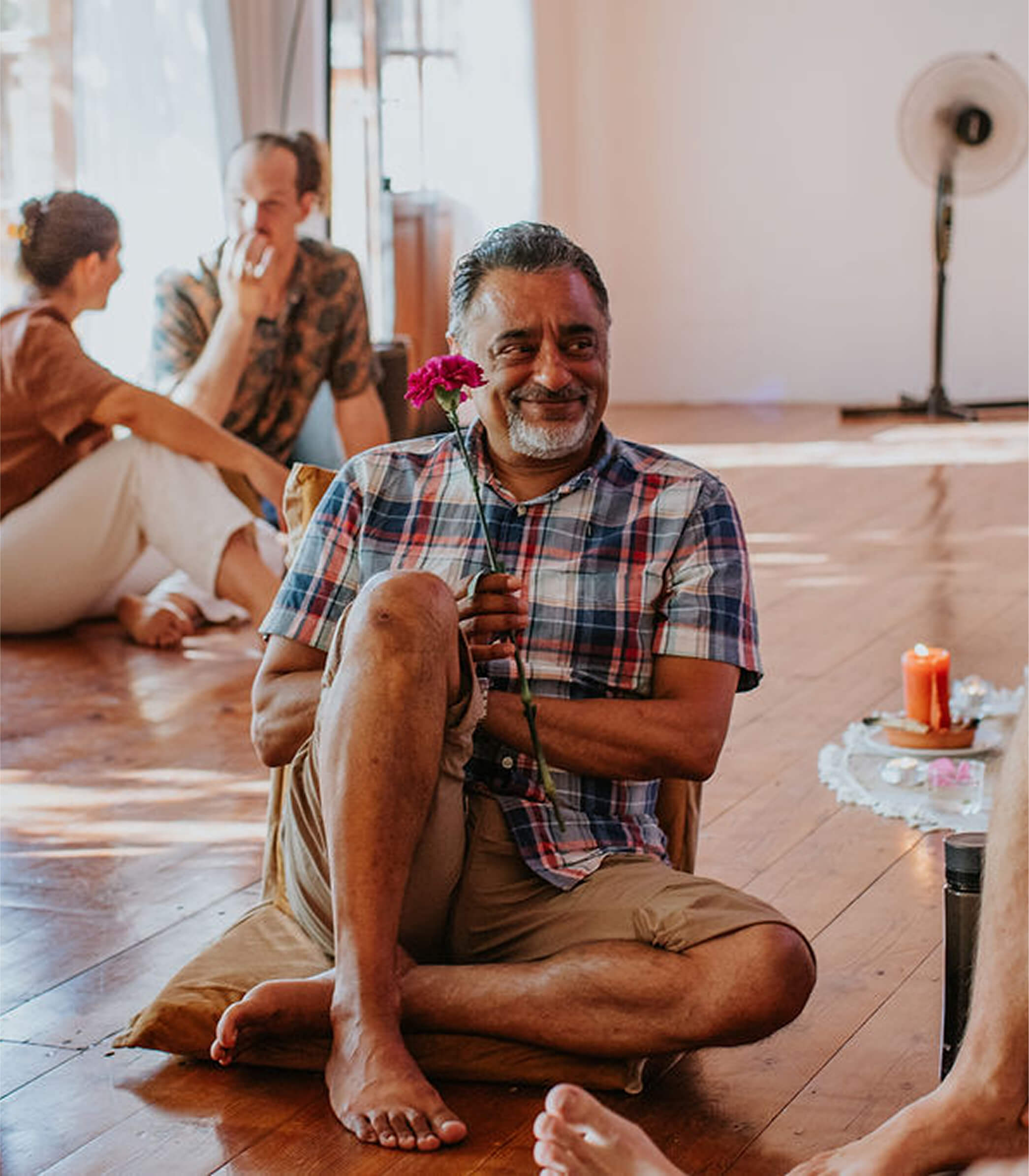 Smiling man in plaid shirt sitting cross-legged on a cushion holding a pink flower, with two people talking in the background on a wooden floor.