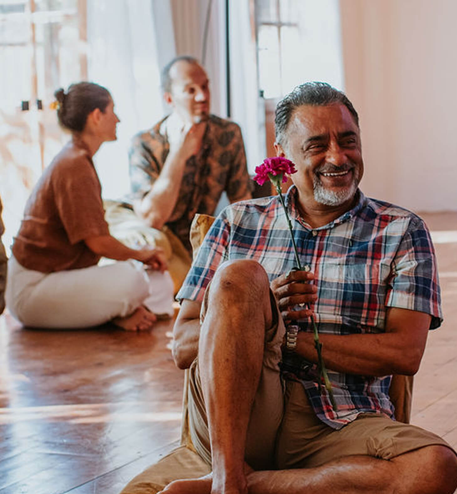 Smiling man in a plaid shirt sitting on the floor holding a pink carnation with two people talking in the background.