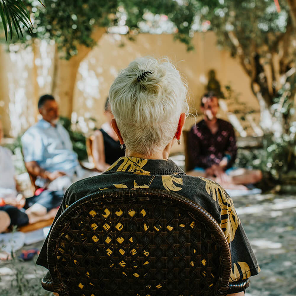Person with short white hair and patterned shirt sitting on a woven chair, facing a small group seated outdoors in shaded garden.