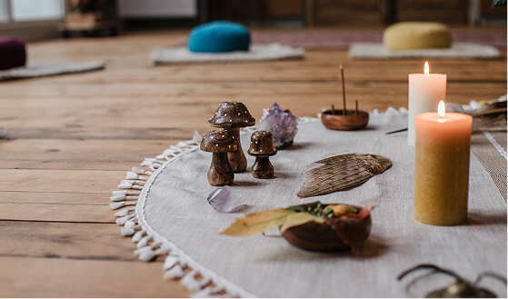 Close-up of a ritual setup on a white fringed cloth with lit candles, wooden mushroom figurines, crystals, and a feather on a wooden floor.