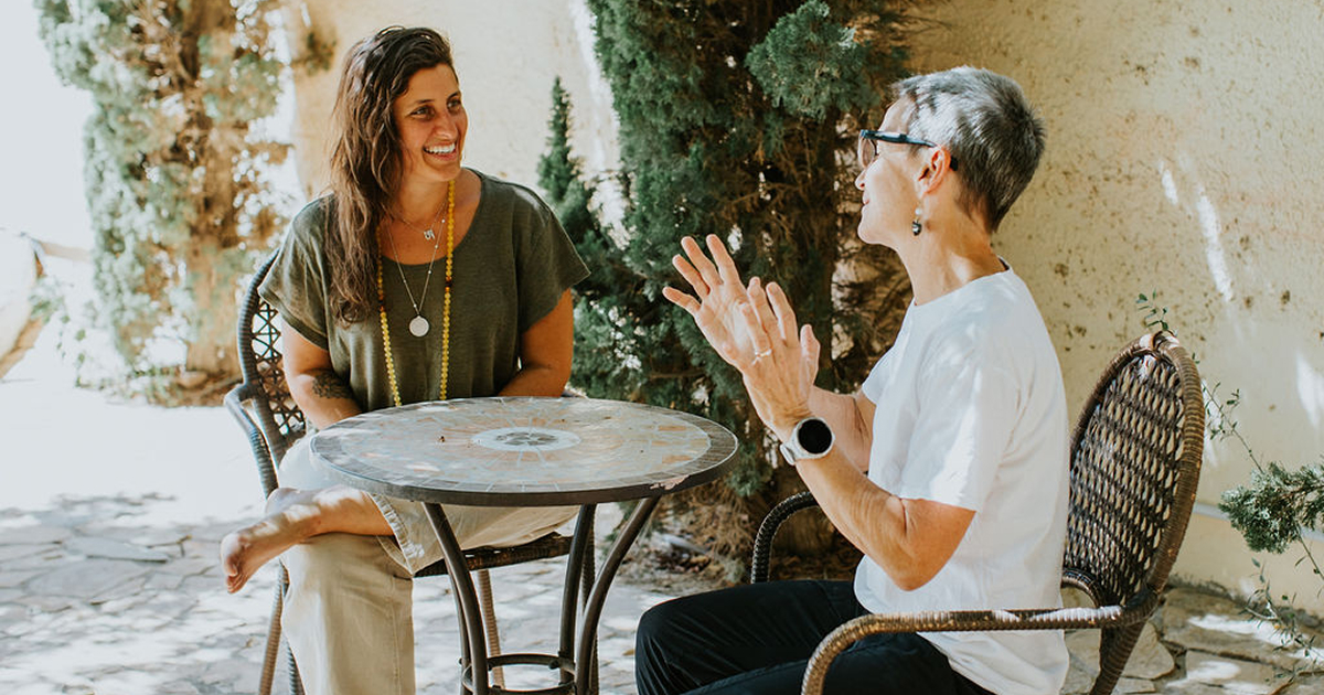 Two women sitting outdoors at a small round table, engaged in a lively conversation with trees and sunlight in the background.