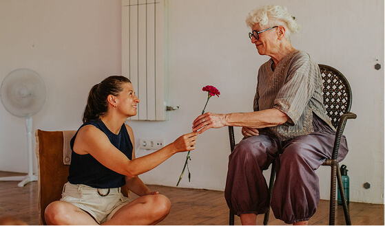 Young woman sitting cross-legged on the floor smiling as she hands a red carnation to an elderly woman seated in a chair.