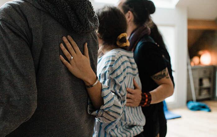 Three people embracing each other indoors, showing a woman's hand with a ring resting on a man's shoulder.