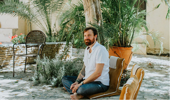 Bearded man in white shirt sitting cross-legged on a cushion outdoors near plants and stone wall, looking to the side.