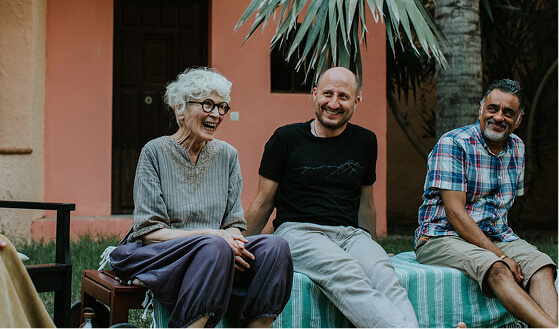 Three people sitting outdoors on a bench and a striped blanket, smiling and chatting near a peach-colored building with palm trees.
