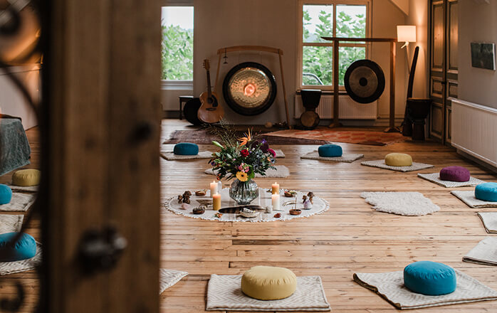 Spacious wooden-floored room set up with colorful meditation cushions, mats, candles, flowers, and gongs for a ceremony or group meditation.