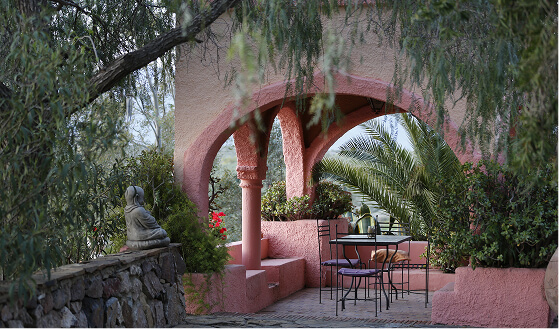 Outdoor seating area with a black metal table and chairs under pink arches surrounded by lush greenery and a stone wall with a Buddha statue.