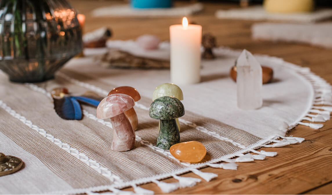 Table with small polished stone mushrooms, a lit white candle, a blue feather, and a clear quartz crystal on a striped cloth.
