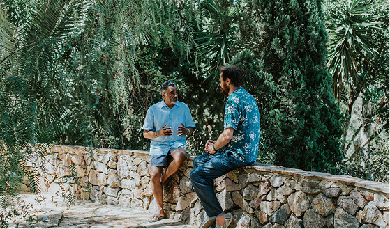 Two men having a conversation while sitting on a low stone wall surrounded by lush greenery.